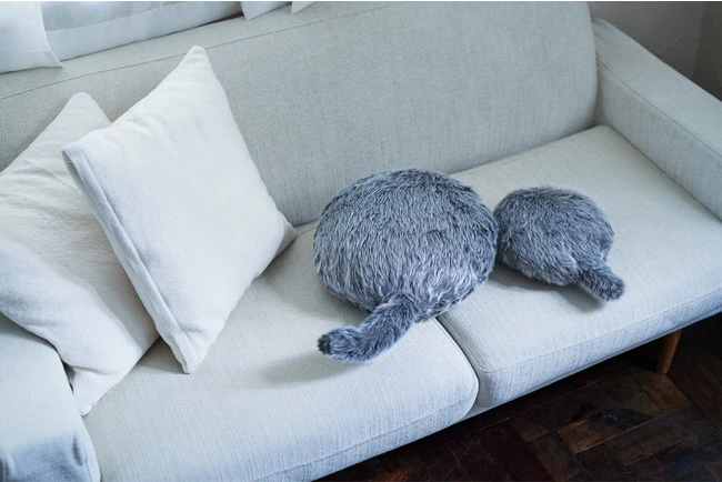 A grey Petit Qoobo robotic cushion with a curved tail on a chair.